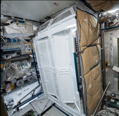 Image of the Universal Waste Management System (UWMS) toilet stall on the International Space Station. The image shows the compact, high-tech toilet unit designed for use in zero-gravity, featuring a vacuum suction system, urine funnel, and waste storage container. The stall is equipped with a raised and tilted seat to enhance comfort and usability for astronauts.